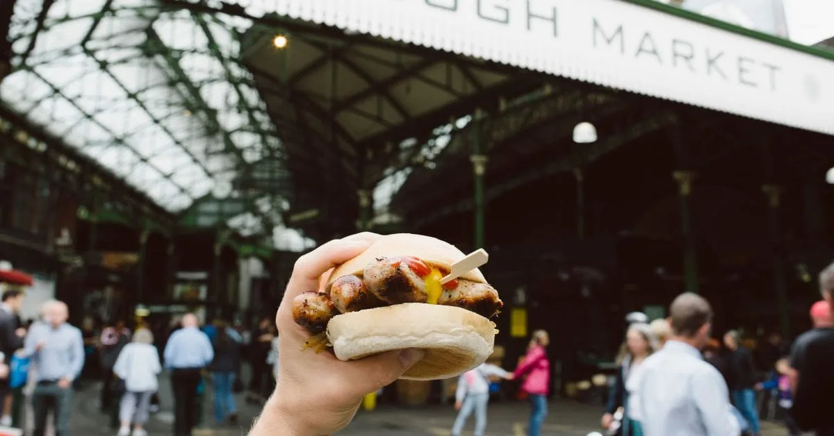 A hand holding a sausage sandwich at Borough Market in London, showcasing the vibrant street food scene